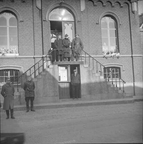 Op het bordes van het gemeentehuis van Aarle-Rixtel zien we vier mensen staan: van links naar rechts een man en een vrouw met hoed, achter hen een burger; rechts staat een hoge militair met helm, met op de punten van zijn boord aan beide kanten drie insignes. Achter het raam lijkt iemand te telefoneren. (Foto: Johan Prinsen, Collectie familie Prinsen, 1939)